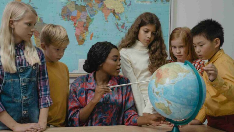 Teacher explaining world map and globe to diverse students in classroom, symbolizing education and future student loan challenges in 2025