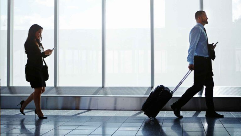 Business travelers walking through an airport, carrying a sleek Goyard travel bag and checking their phones.